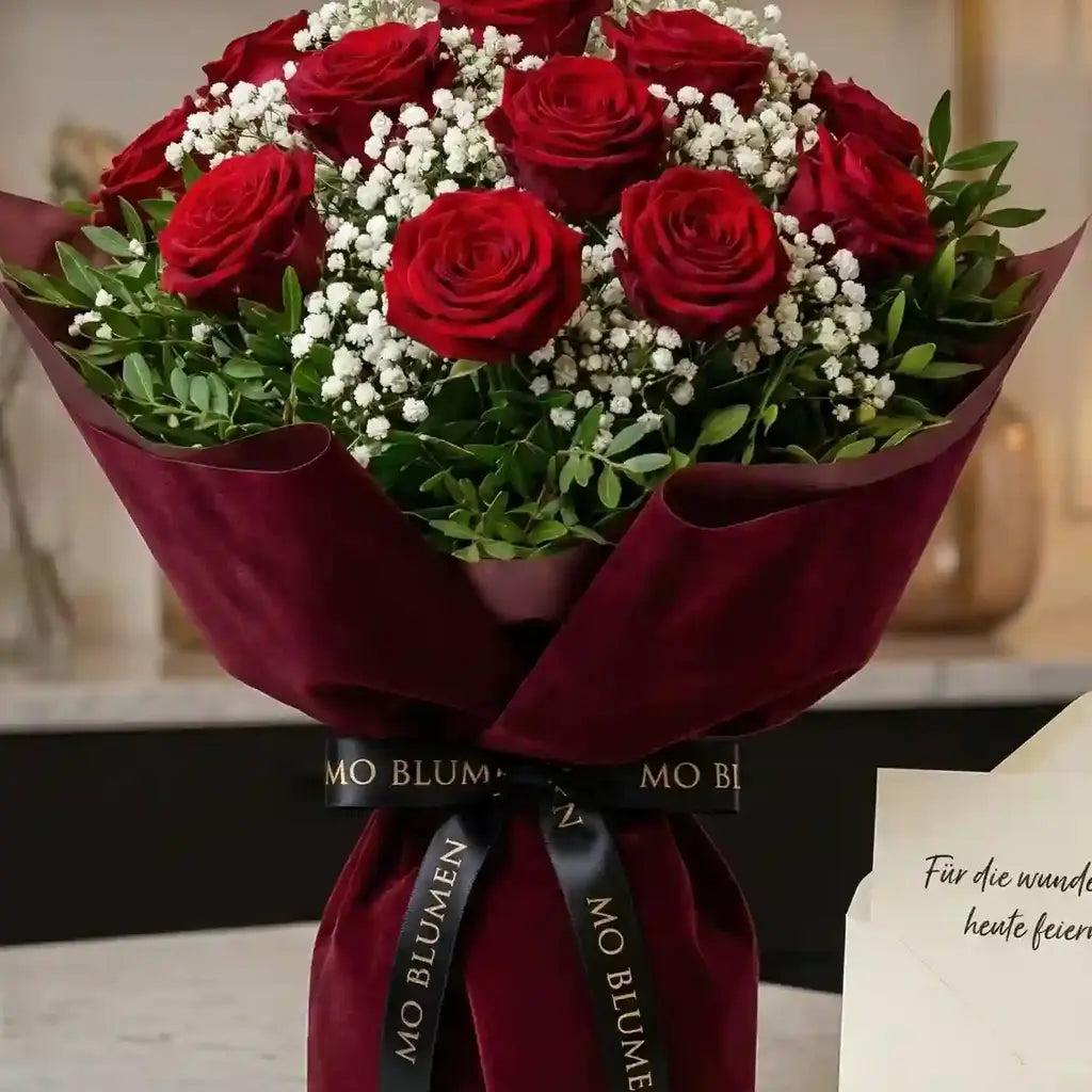 Romantischer Strauß aus roten Rosen von MO BLUMEN, eingewickelt in dunkelrotes Samtpapier, mit Schleierkraut, grünen Blättern und schwarzem Band.